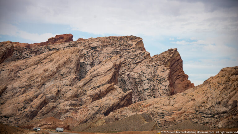 Diagonal sandstone layers rise above a roadside plain with vehicles at their base.