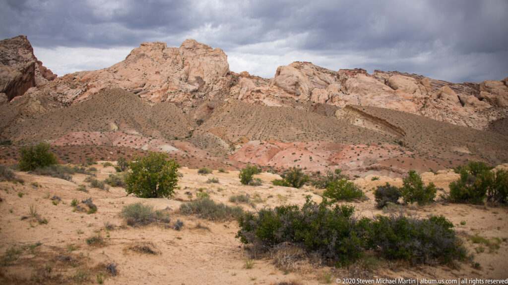 Pale layered ridges rise above desert shrubs beneath a dark, clouded sky.