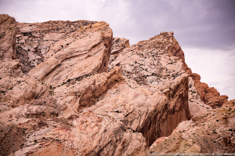 Cream and rust-colored rock beds tilt steeply upward with shrubs in cracks.