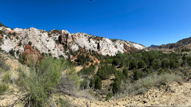 A pale, red-streaked ridge stretches above sagebrush and juniper beneath a blue sky.