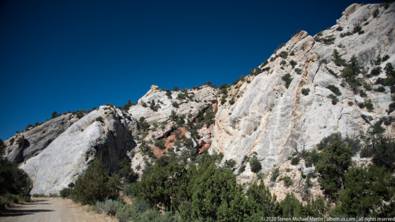 A pale fractured escarpment rises above shrubs, with a dirt road at its base.