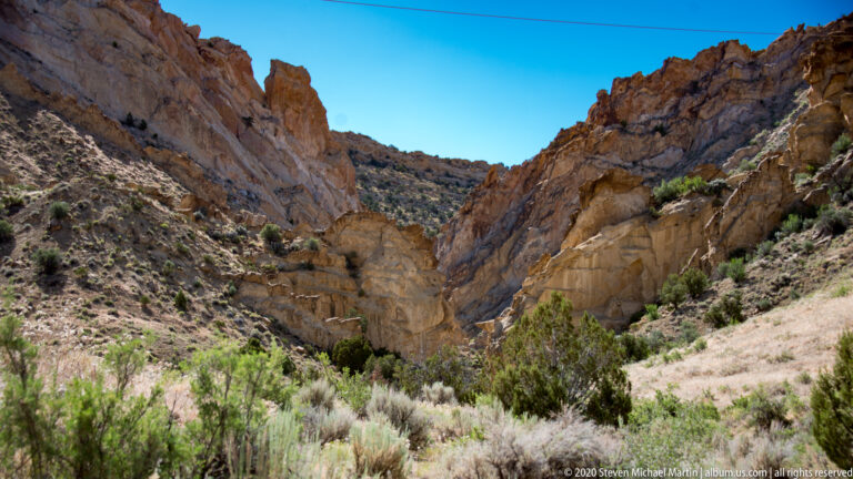 A brush-lined wash leads between converging pale and red cliffs into a narrow canyon.