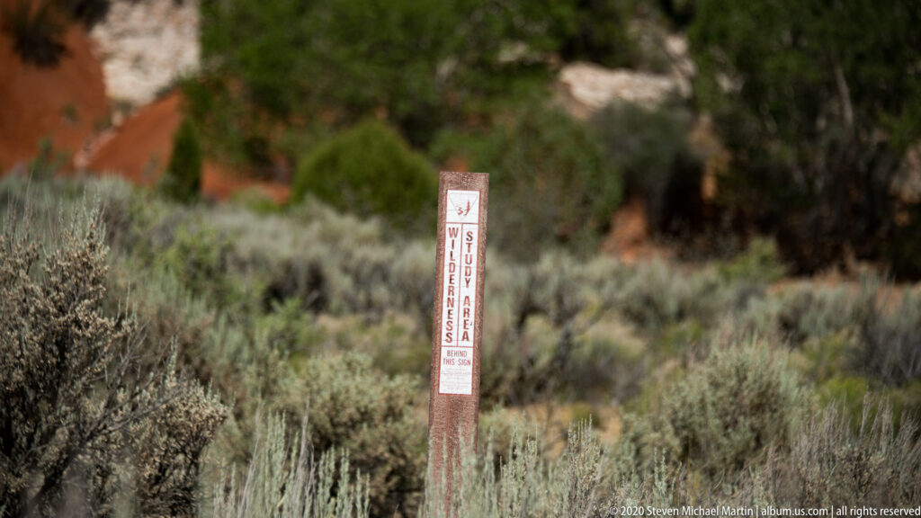A narrow vertical wildlife warning sign stands among sagebrush before blurred red rock and trees.