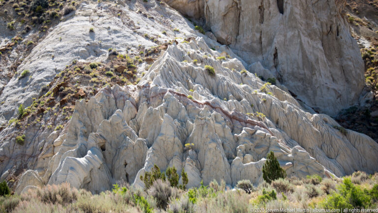 Cracked pale rock ridges descend from a cliff toward shrubs and grasses in a drainage.