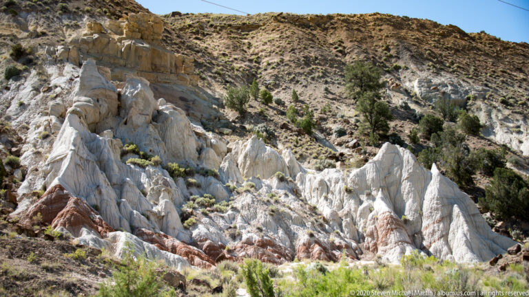 Pale jagged pinnacles rise from a red-and-white hillside above low sagebrush and distant dark mountain slopes.
