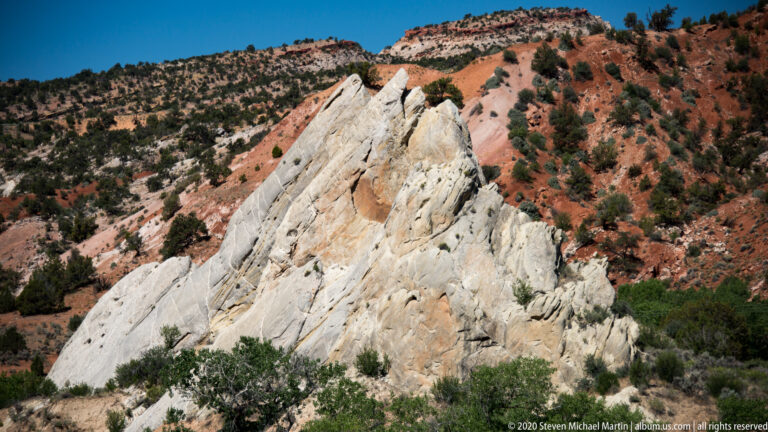 A long pale rock fin crosses a slope of red and white bands above shrubs.
