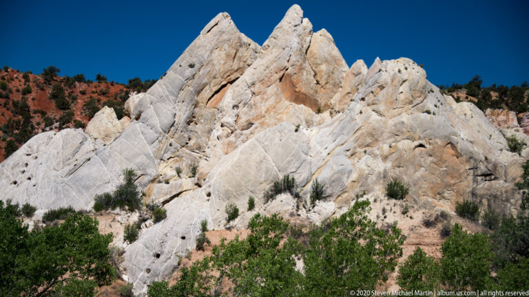 Sharp pale sandstone peaks rise above dark shrubs, with layered red hills behind them.