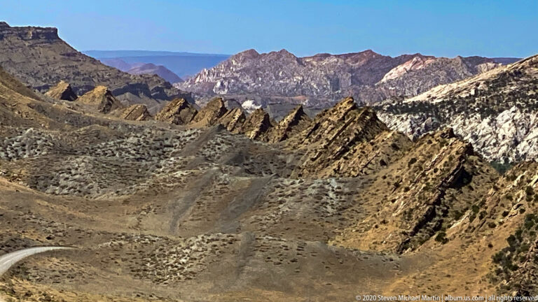 Folded dark ridges cross a high-desert basin above a road bend and distant mountains.