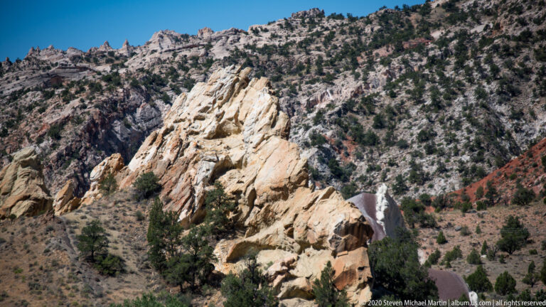 A pale fractured outcrop projects above a ravine with red, cream, and gray slopes.