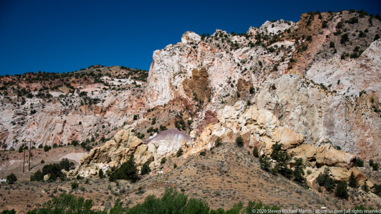 A pale, irregular rock wall dominates the center, with rust-colored streaks, pink rock, and sparse shrubs below.