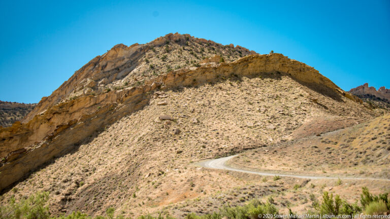 A road forms a tight bend across an open pale slope, with layered rock hills rising behind it.