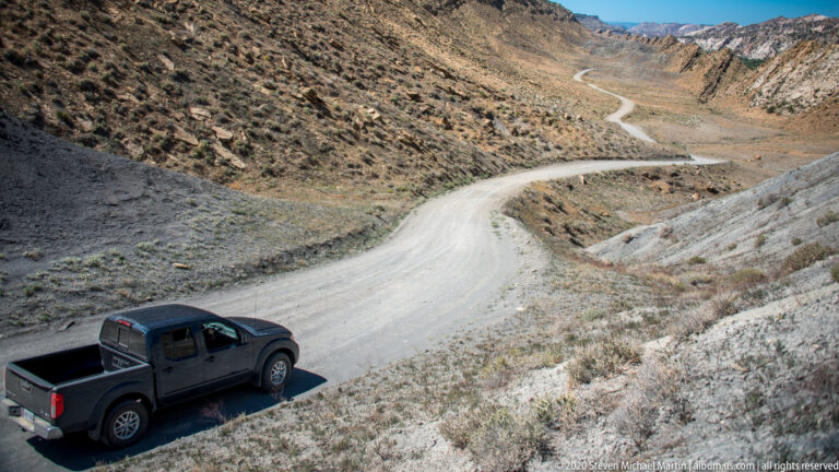 A dark pickup truck is parked at lower left while a pale road snakes uphill between dry slopes.