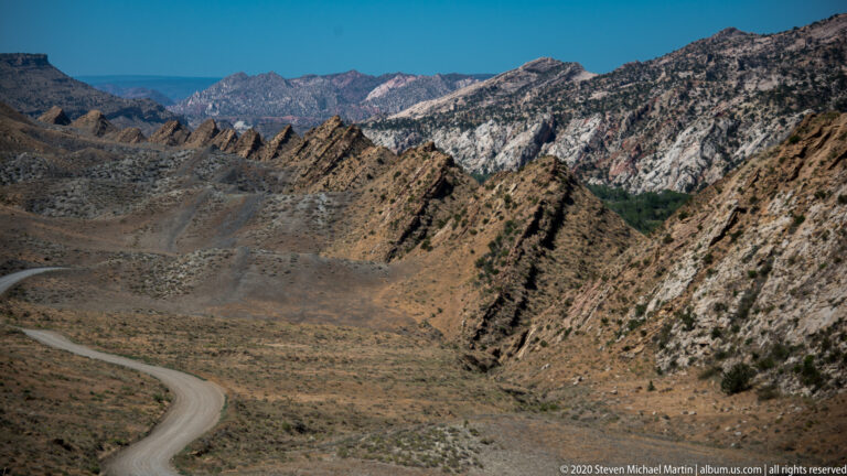 The narrow road follows the lower left slope while a broad tan valley and striped ridge fill the view.