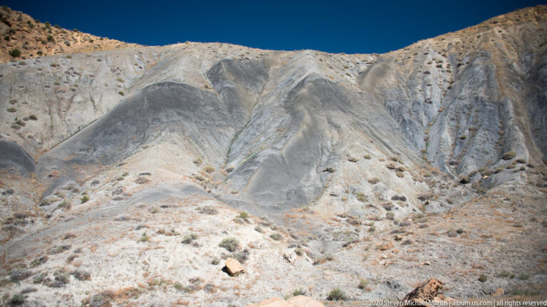 The close slope is scored by branching channels and rounded ridges, with scattered dark stones and small plants.