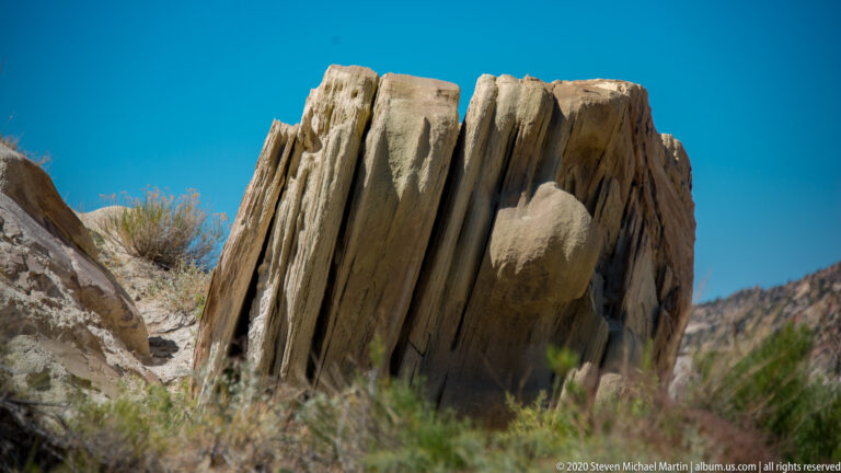 A blocky, vertically fluted rock face stands between low brush and blue sky.
