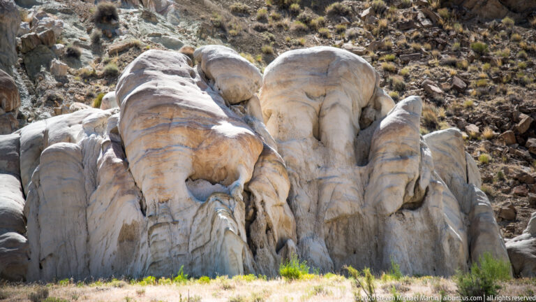 Large smooth pale formations with faint pink bands fill the foreground beneath scattered scrub.