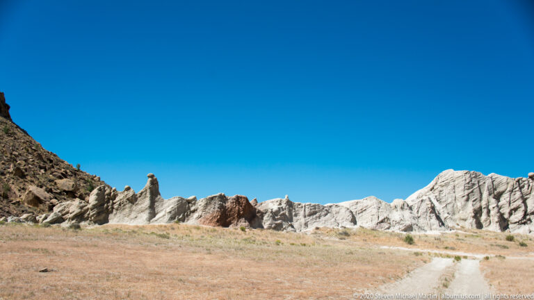The low white and pink rock wall spans the horizon above tawny grassland, with a pale track at right.