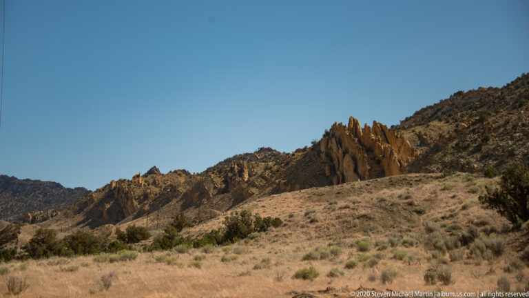 A jagged rust-colored rock ridge rises above pale grasses and dark shrub-covered hills.