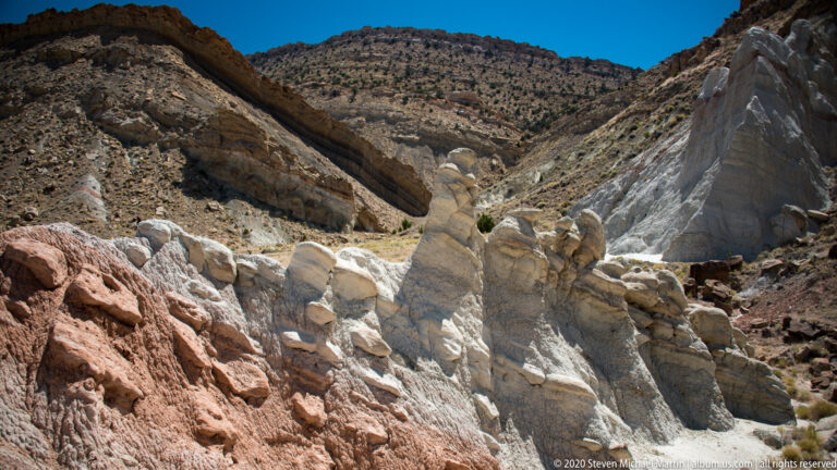 A broad view of pale rock slopes, pink bands, a central wash, and a dark hill beyond.