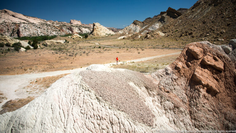 A tiny red-clad figure stands on a light road amid open tan ground and layered hills.
