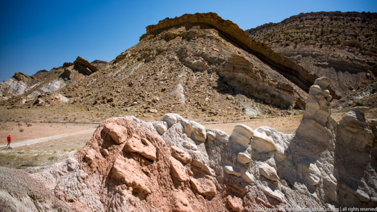 White and pink eroded rock fills the foreground; a small red figure stands beside a pale route below.