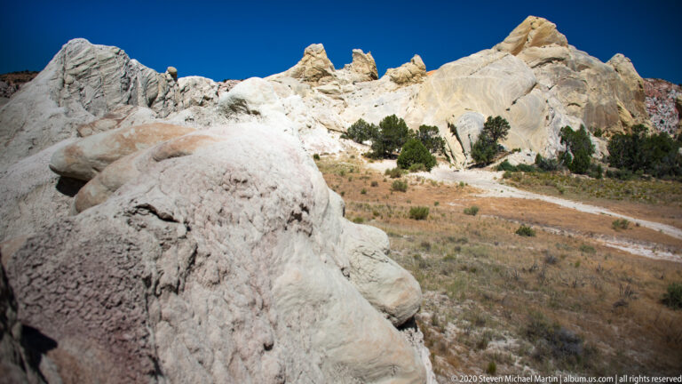 The close boulder dominates the left; pale rock ridges and small evergreens rise across the slope beyond.
