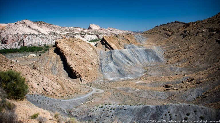 A pale road loops through the lower basin beneath pink and gray slopes and distant white cliffs.