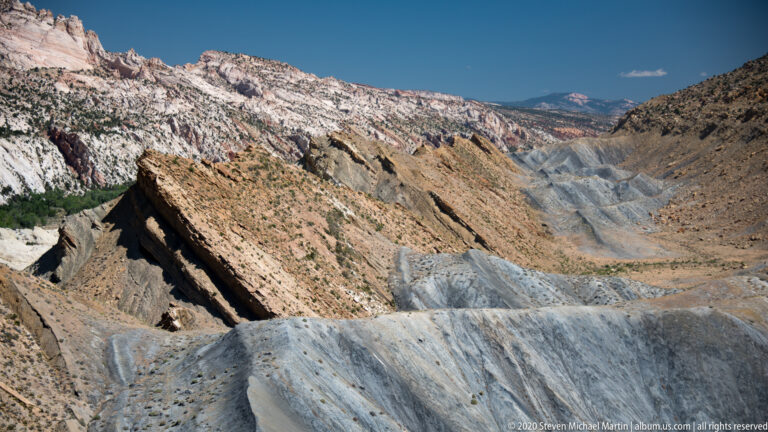 The foreground slope rises toward diagonal tan ridges and a long pale escarpment beneath blue sky.
