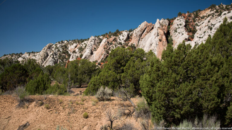 Pale and red rock fins rise above dense juniper and sagebrush on a sandy slope.