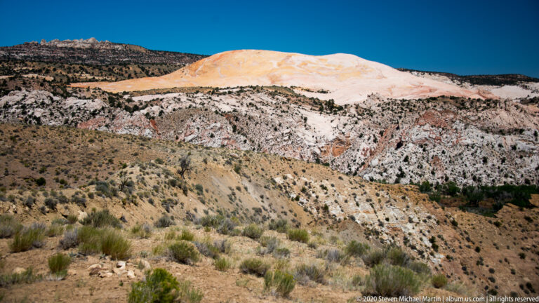 A rounded cream-colored dome rises above white and pink strata and scrub-covered hills.