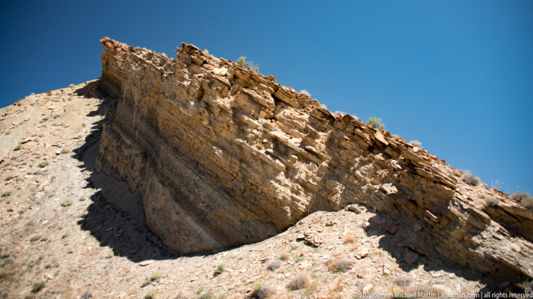 A steep cream sandstone ledge shows fractured layers and angular weathered edges against blue sky.