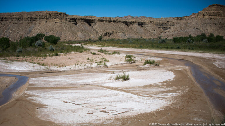 A narrow stream curves through a pale mineral flat below a low layered bluff.