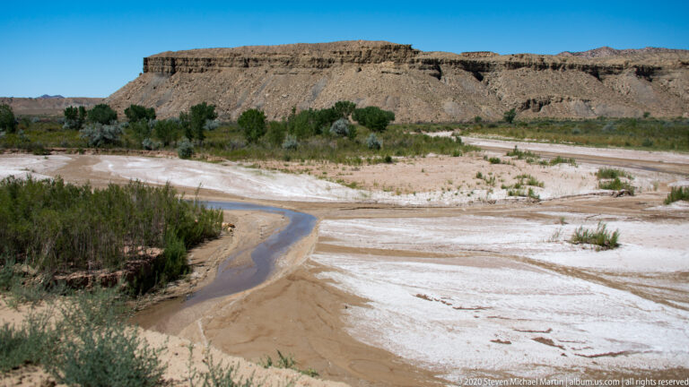 A winding dark stream cuts through a pale wash bordered by brush and low cliffs.