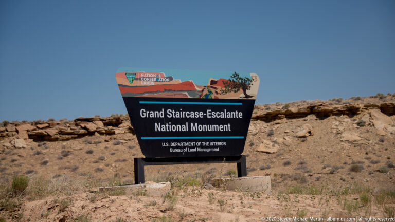 A sign identifies Grand Staircase-Escalante National Monument above a low stone wall in desert scrub.