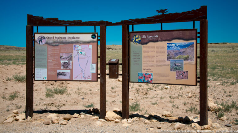 Two timber-framed interpretive panels stand beneath a canopy in a rocky desert setting.