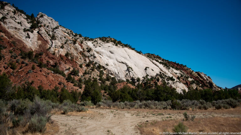 A pale red-streaked cliff rises above a sandy clearing bordered by sagebrush and juniper.