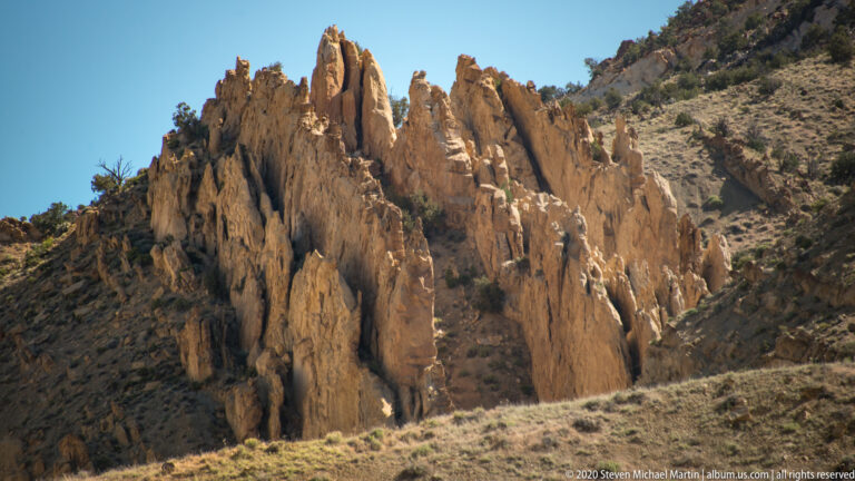 Tall ochre sandstone fins rise together above grasses and shrubs on a steep hillside.