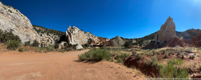A narrow sandstone spire rises beside a winding dirt road among pale cliffs and red ridges.