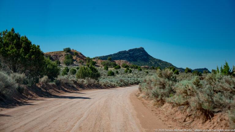 A sandy road curves through sagebrush toward a dark, shrub-covered ridge beneath blue sky.