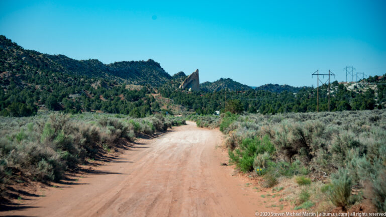 A dirt road passes through shrubs toward a pointed sandstone formation, with power lines on the right.