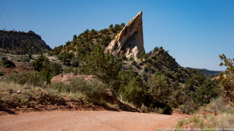 A tall pale sandstone fin with red streaks rises above shrubs and trees beside a dirt road.
