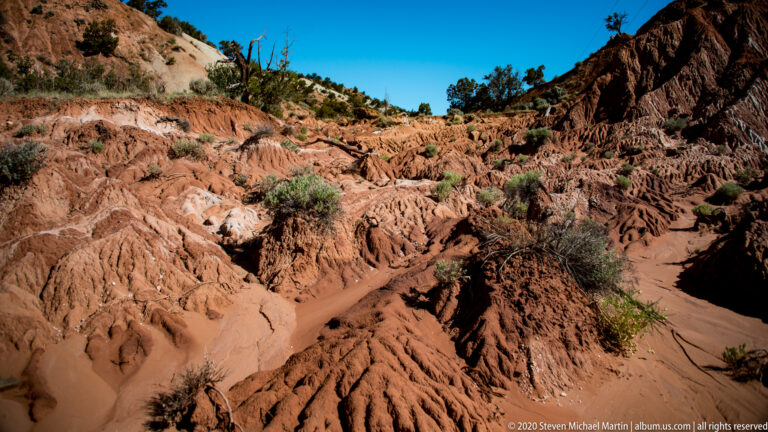 A sandy wash runs between ribbed red-rock slopes and a dark cliff in a deeply eroded basin.