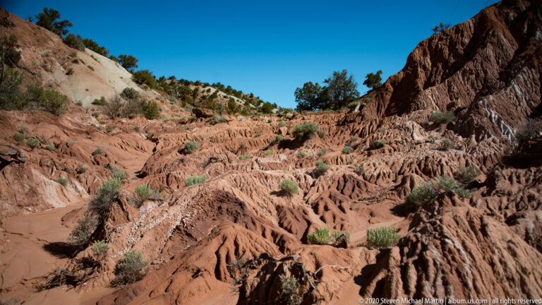 Ribbed red sandstone ridges surround a pale wash between a dark cliff and light slope.