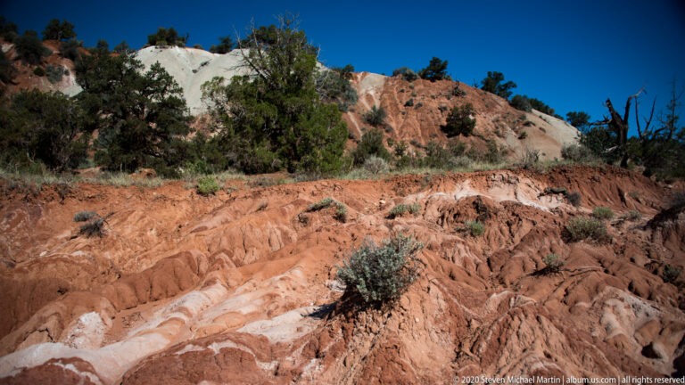 Cream and red rock bands slope through a shrub-dotted hillside above a pale wash.