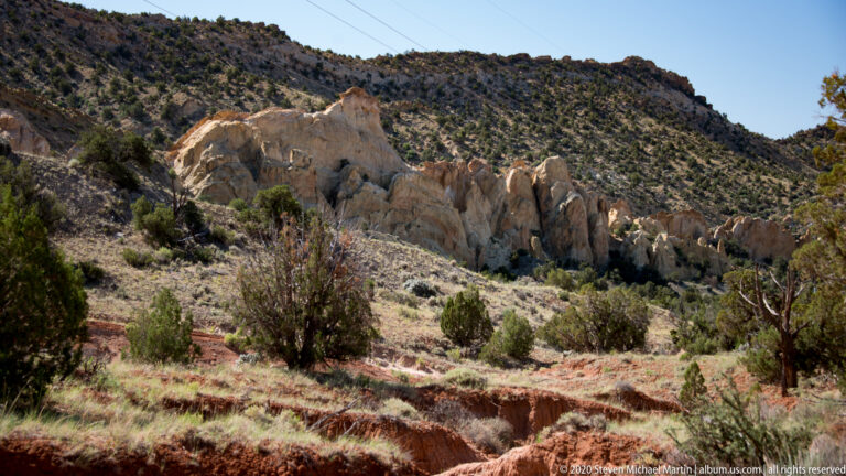 A pale fractured outcrop rises above sagebrush and a red wash, crossed by thin wires.