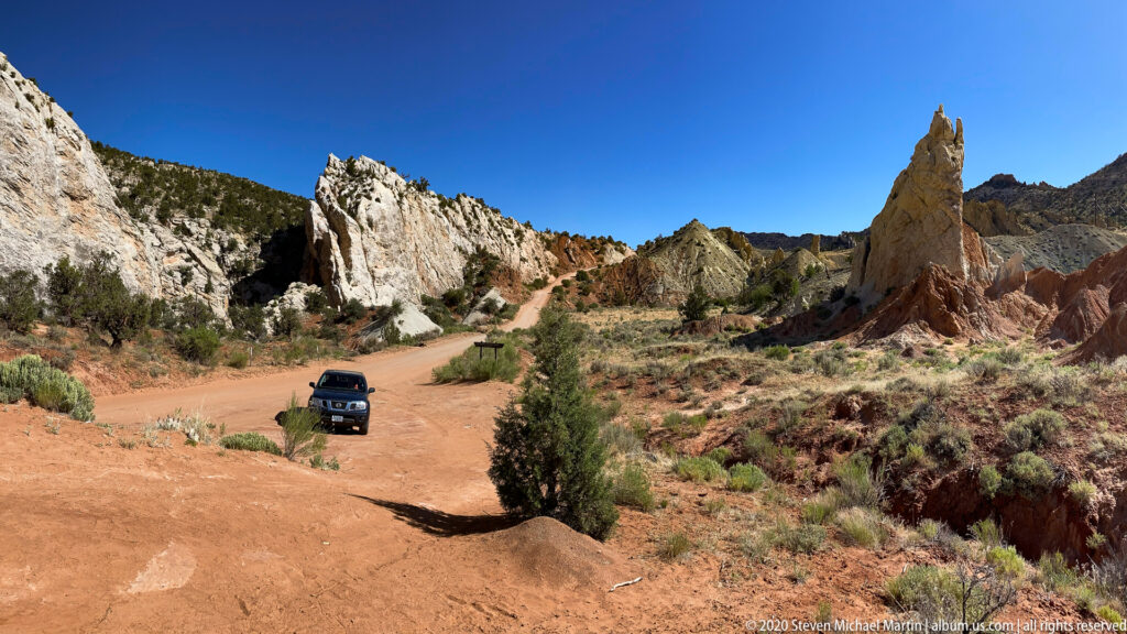 An SUV drives along a sandy track toward pale cliffs and a tall red rock spire.