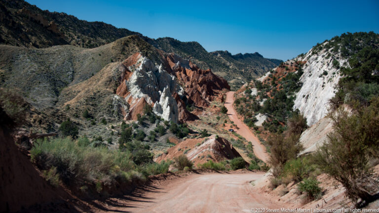 A dirt road descends between pale and rust-red canyon walls toward a hidden bend.