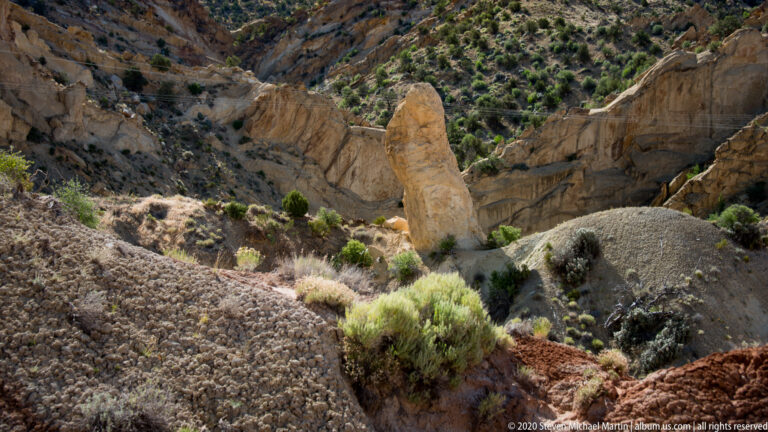 A narrow tan sandstone pillar stands among pale boulders and low shrubs below fractured cliffs.