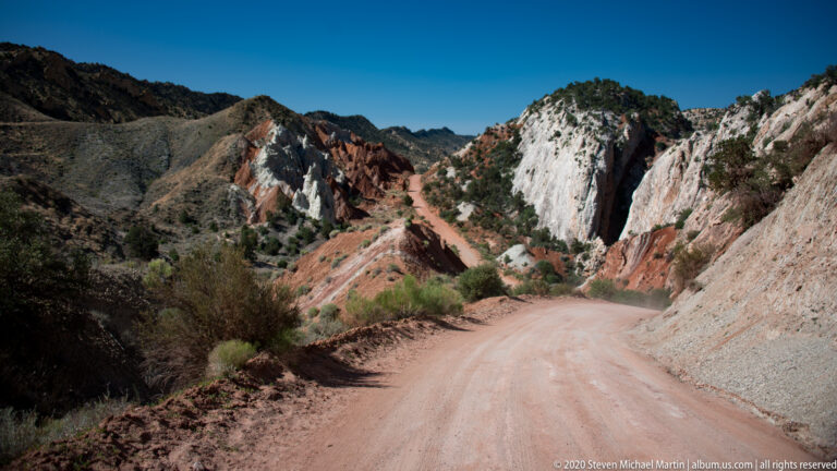 A dirt road curves between a dark red ridge and pale cliff toward striped formations.