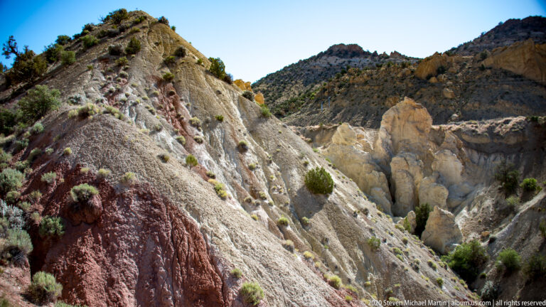 Pale sandstone with gray and red bands rises above red ledges and scattered shrubs.